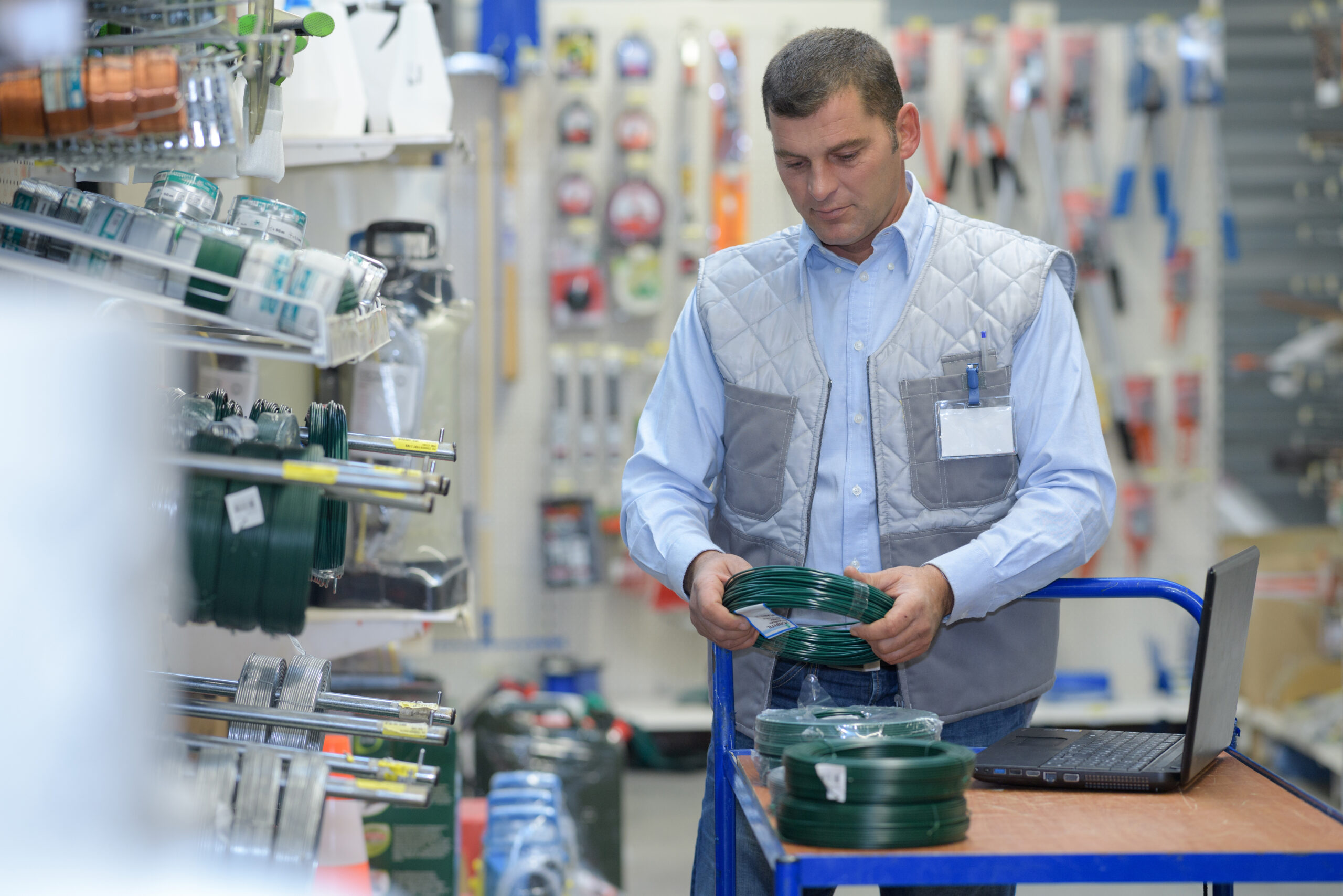 Hardware store employee organizing inventory with a laptop on a cart, symbolizing inventory management and optimization.